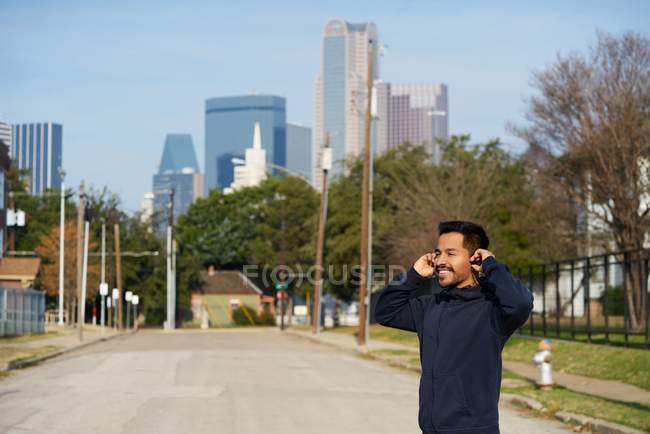Zufriedener hispanischer männlicher Athlet lächelt, während er beim morgendlichen Joggen in der Innenstadt Kopfhörer einsetzt — Stockfoto