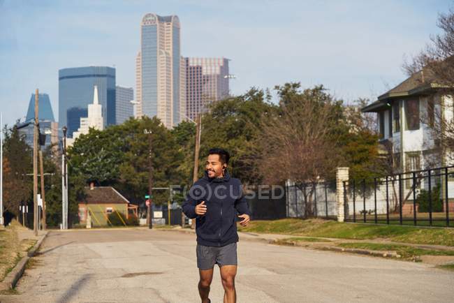 Happy Hispanic male athlete in sportswear with headphones running along empty road at downtown in Dallas — Stock Photo