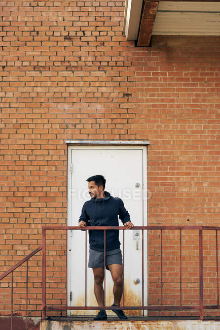 Hombre joven hispano en activo de pie y mirando hacia otro lado en el balcón con puerta blanca cerrada y pared de ladrillo - foto de stock