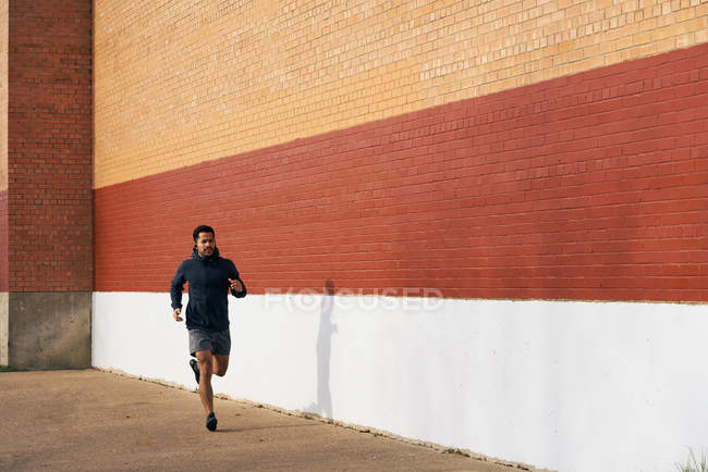 Vista lateral del hombre deportivo hispano en forma en ropa activa escuchando música mientras trota por la calle de la ciudad en Dallas - foto de stock