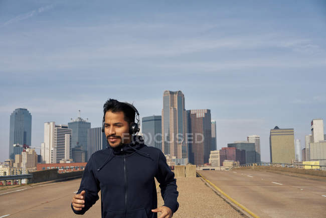 Jogador masculino hispânico com capuz casual usando fones de ouvido enquanto corre com céu azul acima do centro da cidade em Dallas, Texas — Fotografia de Stock