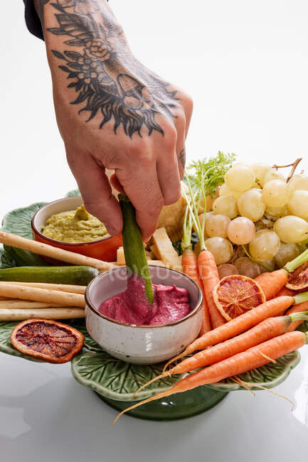 Brazo tatuado sumergiendo pepino en salsa en el plato con verduras - foto de stock