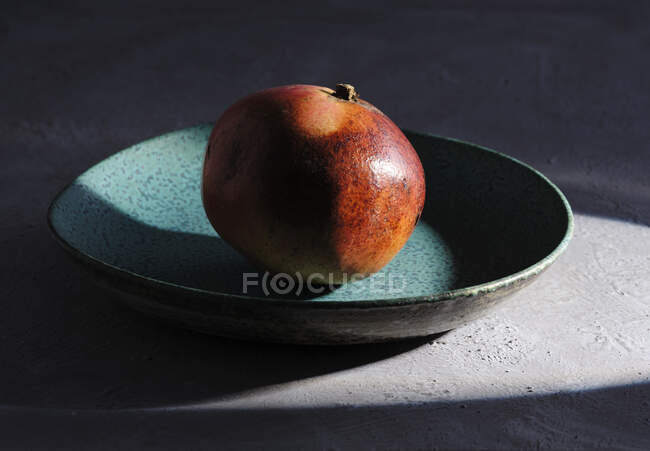 Blue plate with single ripe fresh pomegranate on concrete surface lit by sunbeam — Stock Photo