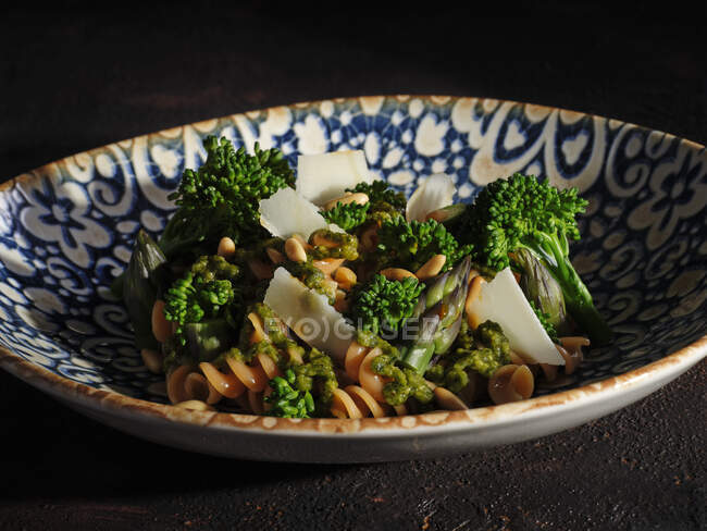 Plate of pasta with broccoli and pine nuts garnished with thinly sliced cheese on table — Stock Photo