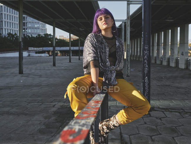 Stylish woman with bright purple hairstyle in yellow pants sitting on metal fence in city station, looking in camera — Stock Photo