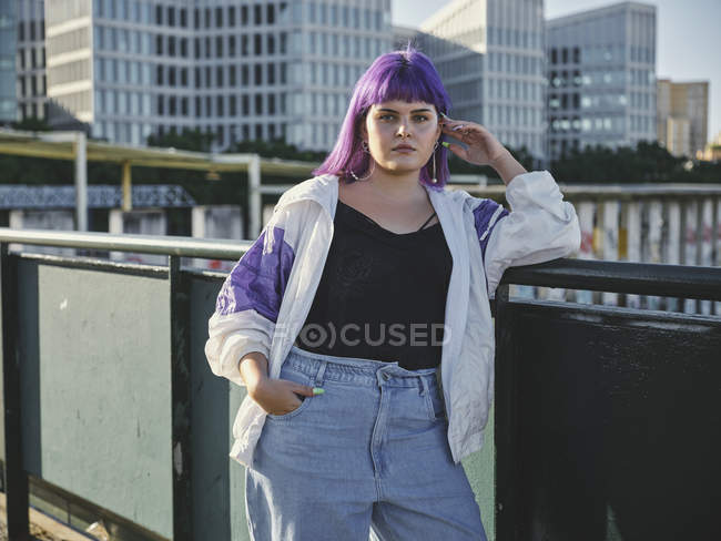 Модная стильная женщина с фиолетовой прической, касающаяся лица и опирающаяся на металлический забор в центре города — стоковое фото