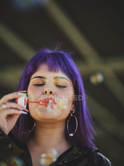 Jolie femme aux cheveux violets soufflant des bulles tenant bouteille avec les yeux fermés avec la main soignée — Photo de stock