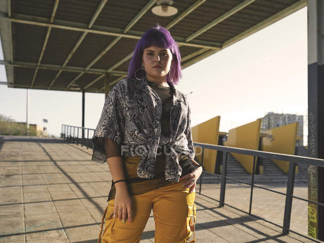 Stylische Frau mit lila Frisur in gelben Hosen steht auf der Straße und schaut in die Kamera — Stockfoto