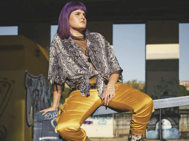 Femme élégante avec une coiffure violette lumineuse en pantalon jaune assis sur une clôture en métal dans la gare de la ville, détournant les yeux — Photo de stock