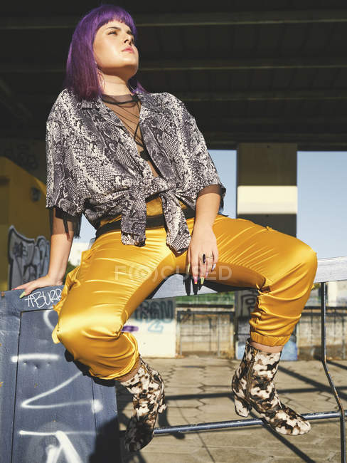 Стильна жінка з яскраво-фіолетовою зачіскою в жовтих штанях, що сидять на металевому паркані на міській станції, дивлячись подалі — стокове фото
