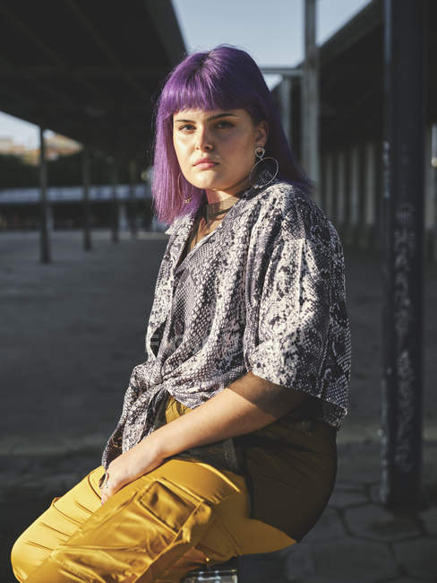 Стильна жінка з яскраво-фіолетовою зачіскою в жовтих штанях, що сидять на металевому паркані на міській станції, дивлячись в камеру — стокове фото