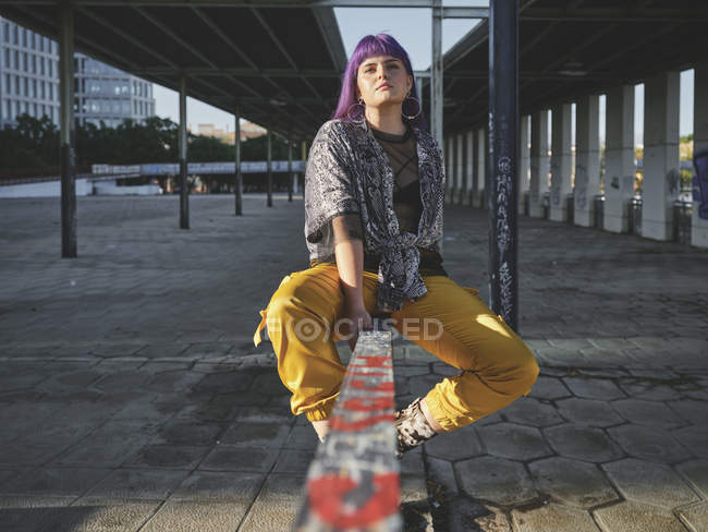 Стильна жінка з яскраво-пурпуровою зачіскою в жовтих штанах сидячи на металевому паркані на міській станції. — стокове фото