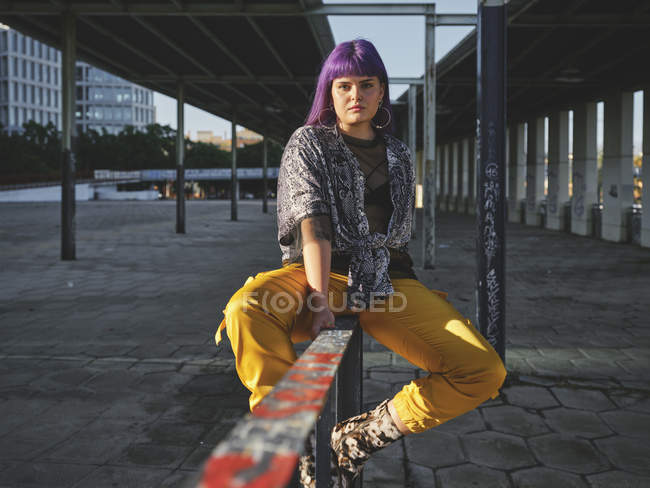 Mulher elegante com penteado roxo brilhante em calças amarelas sentado na cerca de metal na estação da cidade, olhando na câmera — Fotografia de Stock