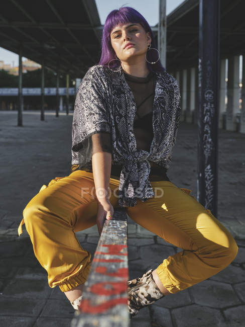 Стильная женщина с ярко-фиолетовой прической в желтых штанах сидит на металлическом заборе на городском вокзале, смотрит в камеру — стоковое фото