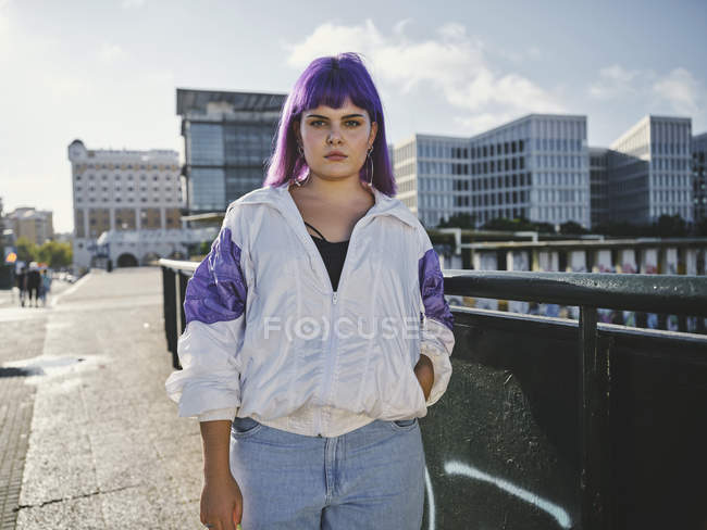 Стильна жінка з фіолетовою зачіскою в модному одязі позує в міському мосту в місті — стокове фото