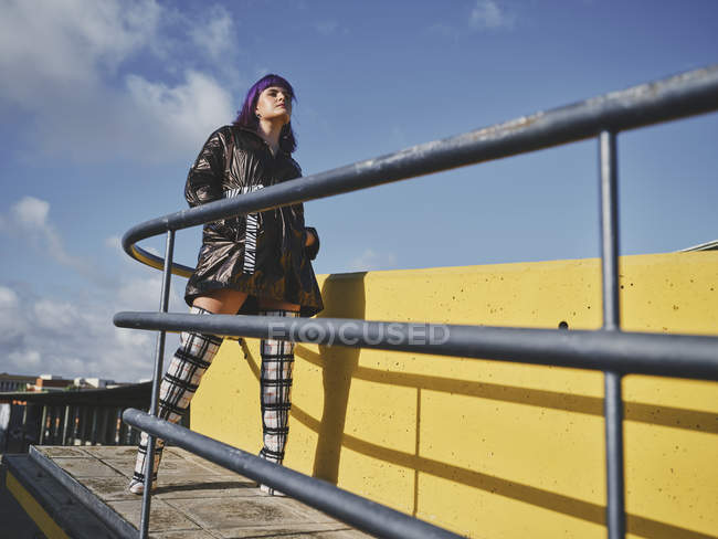 Mulher confiante com penteado roxo em casaco preto brilhante olhando para longe no ponto de vista da cidade com cerca de metal — Fotografia de Stock