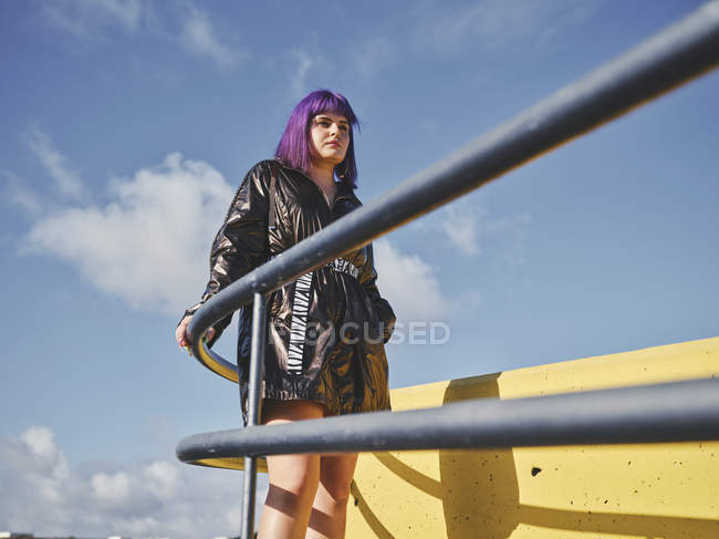 Уверенная женщина с фиолетовой прической в блестящей черной куртке, смотрящая на город с металлическим забором — стоковое фото