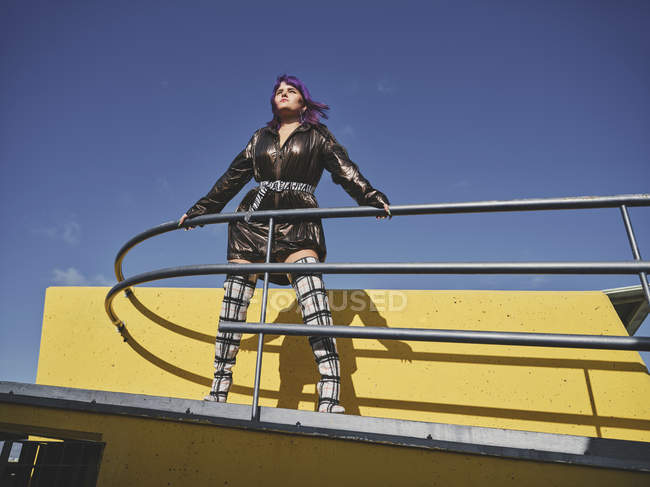 Basso angolo di donna con acconciatura viola in giacca nera lucida guardando lontano sul punto di vista della città contro il cielo blu — Foto stock
