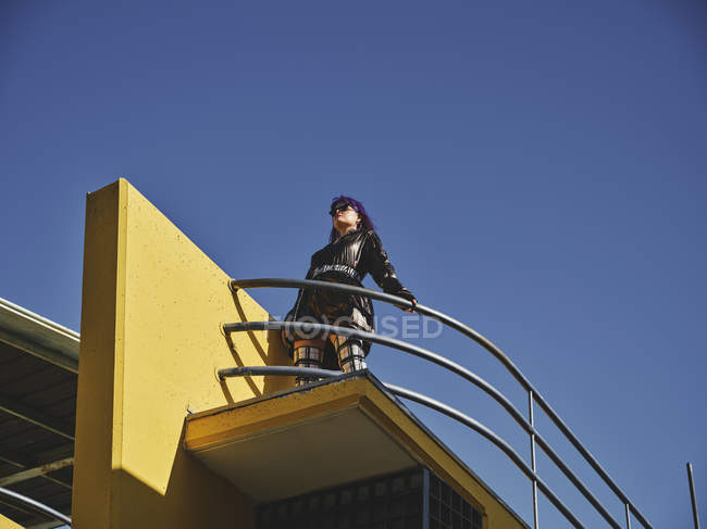 Низький кут жінки з фіолетовою зачіскою в блискучій чорній куртці, що дивиться на точку зору міста на блакитне небо — стокове фото