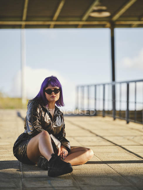 Mujer joven en gafas de sol con peinado morado en chaqueta negra brillante cómodamente sentada sobre asfalto con piernas cruzadas en la ciudad - foto de stock
