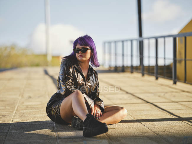 Junge Frau mit Sonnenbrille und lila Frisur in glänzender schwarzer Jacke sitzt gemütlich mit überkreuzten Beinen auf Asphalt in der Stadt — Stockfoto