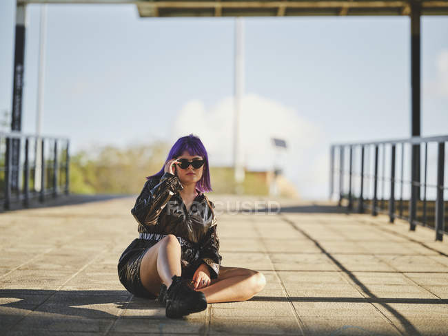 Jeune femme en lunettes de soleil avec coiffure violette en veste noire brillante confortablement assise sur asphalte avec jambes croisées en ville — Photo de stock