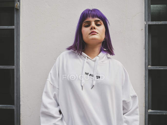 Модна жінка з фіолетовою зачіскою, спираючись на білу стіну і задумливо дивлячись в камеру — стокове фото