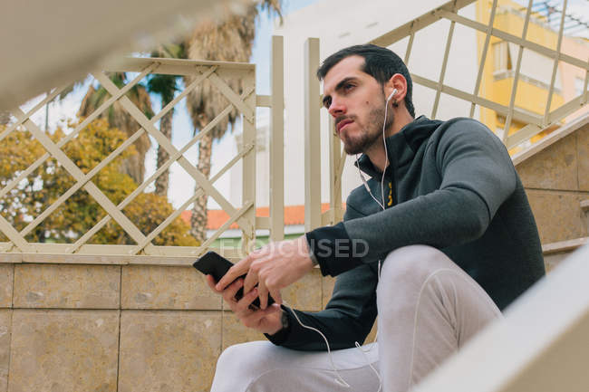 Von unten ein nachdenklicher junger Mann im Trainingsanzug mit Kopfhörer-Nachrichten auf dem Handy, der auf der Treppe sitzt und wegschaut — Stockfoto