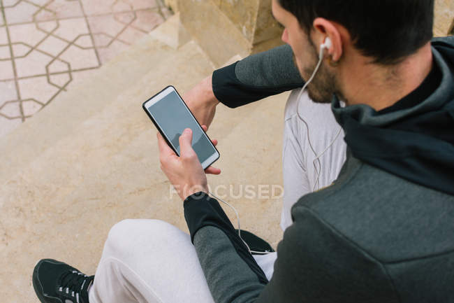 Von oben ein nachdenklicher junger Mann im Trainingsanzug mit Kopfhörer-Nachrichten auf dem Handy, der auf der Treppe sitzend Pause macht — Stockfoto