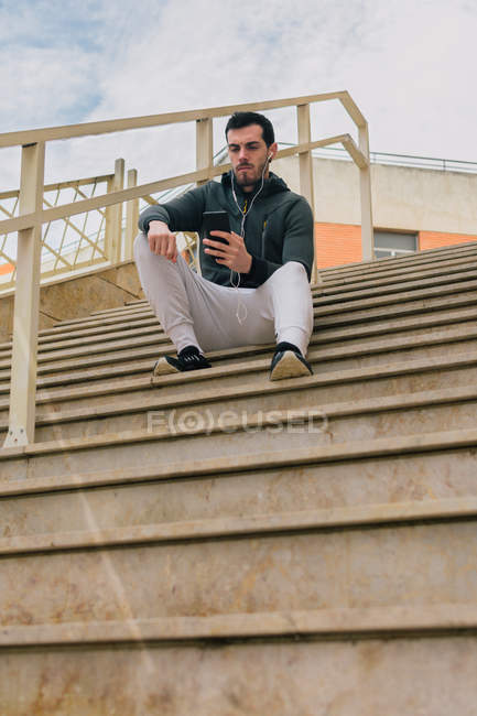 Desde abajo de un joven pensativo en chándal con auriculares mensajería en el teléfono móvil tomando un descanso sentado en las escaleras - foto de stock