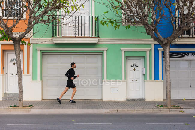 Vista lateral del chico activo en ropa deportiva con auriculares con teléfono en las piernas de entrenamiento de la mano corriendo en la calle urbana - foto de stock