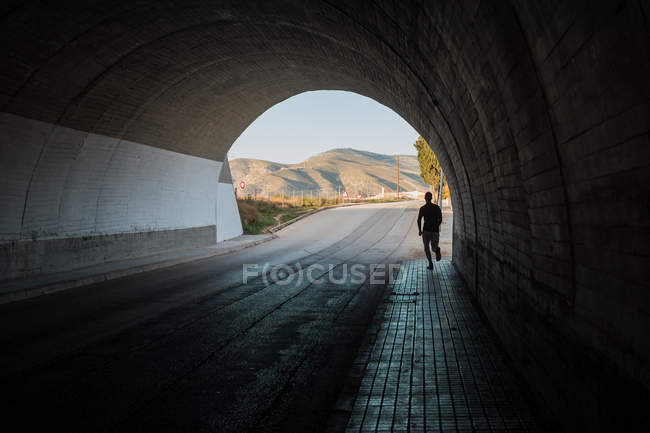 Strong man doing sport exercise working on legs jogging on sidewalk in tunnel on sunny day — Stock Photo