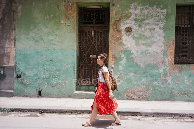 Vista lateral de la hembra de vacaciones en falda roja colorida con mochila y cámara fotográfica caminando a lo largo de la carretera con el edificio viejo en el fondo en Cuba - foto de stock