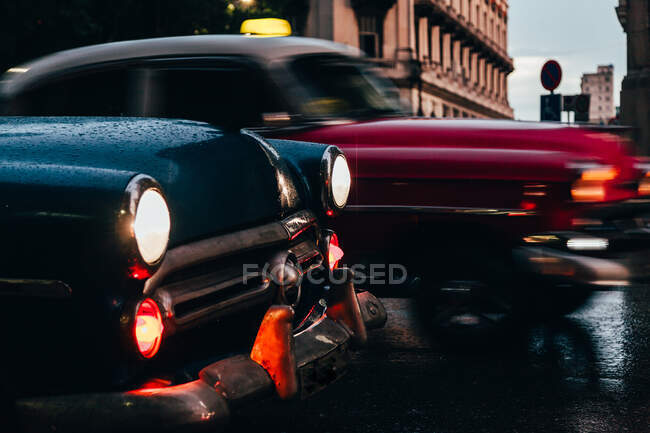Capucha de coche vintage azul con luces encendidas y coche viejo rojo en movimiento sobre fondo sobre tiempo lluvioso en Cuba - foto de stock