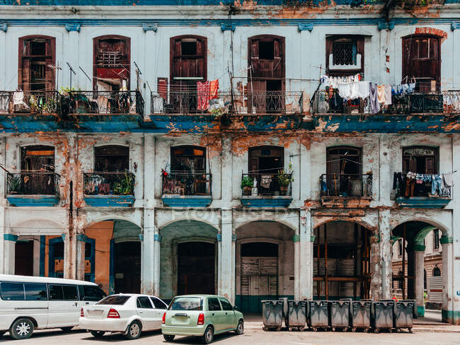 Дилапідований синій будинок з великими вікнами і балконами біля парковок з автомобілями і сміттєвозом на Кубі. — стокове фото