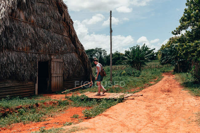 Senhora descansando em roupas casuais elegantes em pé na porta da grande casa rural com telhado de palha e céu azul no fundo em Cuba — Fotografia de Stock