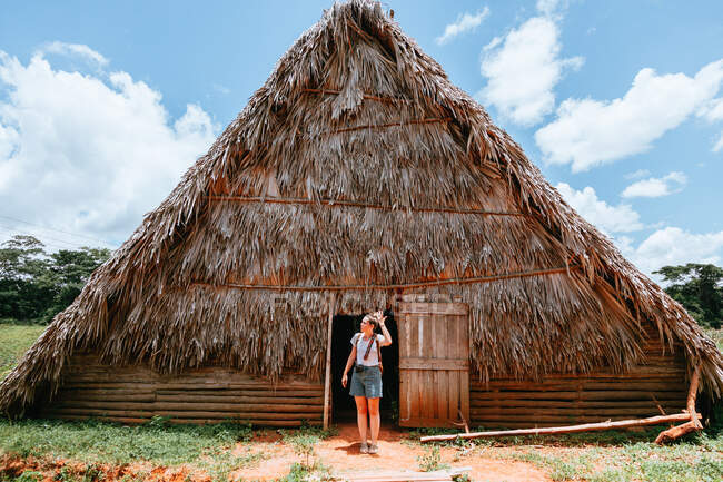 Отдыхающая леди в стильной повседневной одежде стоит в дверях большого сельского дома с соломенной крышей и голубым небом на заднем плане на Кубе — стоковое фото