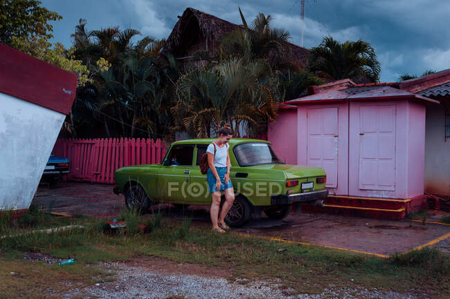 Jovencita en ropa casual riéndose de pie cerca de un viejo auto verde entre casas rurales y plantas tropicales en Cuba - foto de stock