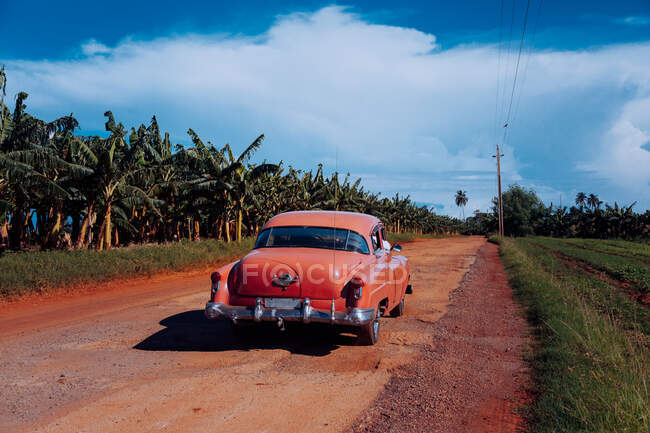 Estrada vermelha de sujeira arenosa com carro antigo vintage com plantas verdes nas laterais e céu cinza nublado no fundo em Cuba — Fotografia de Stock