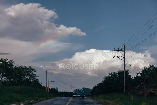 Carretera de asfalto estrecha con coche viejo con plantas verdes en los lados y cielo nublado gris en el fondo en Cuba - foto de stock