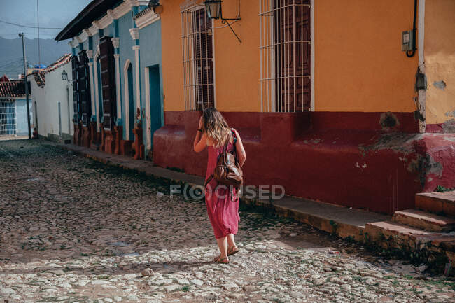 Vista trasera de una mujer sin rostro de vacaciones en vestido rosa claro y mochila caminando por un camino empedrado vacío entre viejos edificios coloridos en Cuba - foto de stock