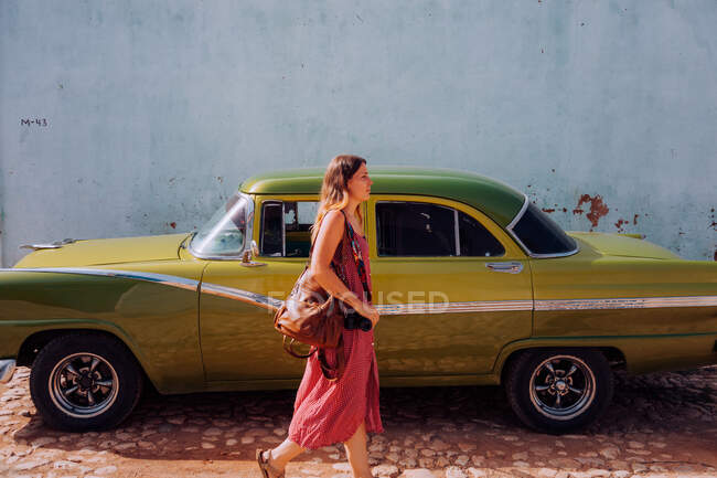Side view of woman on vacation in light pink gown and backpack walking on road with vintage old car parked in Cuba — Stock Photo