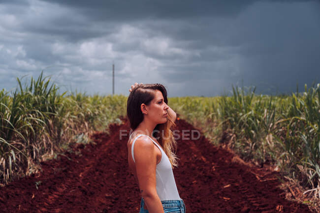 Vista laterale del viaggiatore femminile in abbigliamento casual in piedi vicino al suolo marrone tra le piante tropicali verdi sotto il cielo grigio nuvoloso a Cuba — Foto stock