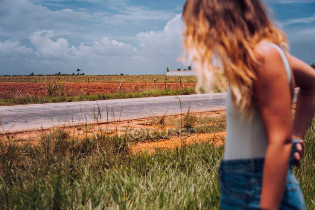 Vista lateral do viajante feminino em desgaste casual em pé na encruzilhada com solo marrom entre as plantas tropicais verdes sob céu nublado cinza em Cuba — Fotografia de Stock