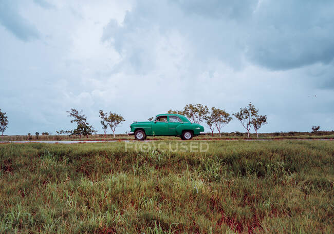 Chemin de terre sablonneux rouge avec vieille voiture vintage avec des plantes vertes sur les côtés et ciel gris nuageux sur le fond à Cuba — Photo de stock