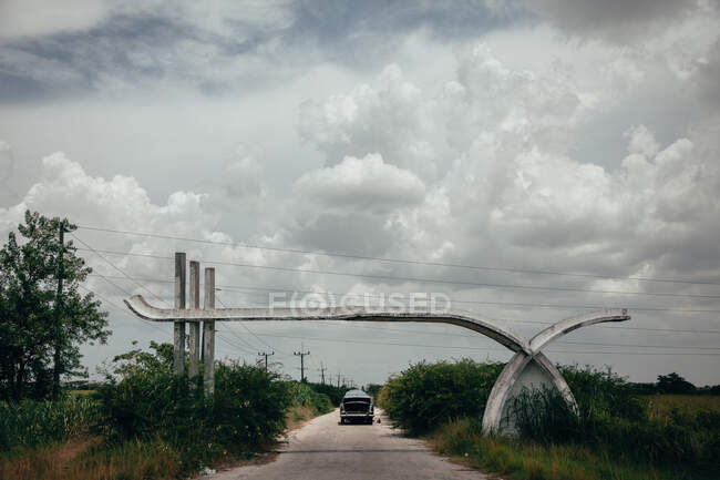 Вузька асфальтна дорога зі старою машиною серед творчої скелястої структури з зеленими рослинами збоку і сірим хмарним небом на задньому плані на Кубі. — стокове фото