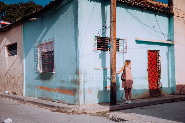 Mujer de vacaciones en vestido ligero y mochila caminando por un camino empedrado vacío entre viejos edificios coloridos en Cuba - foto de stock