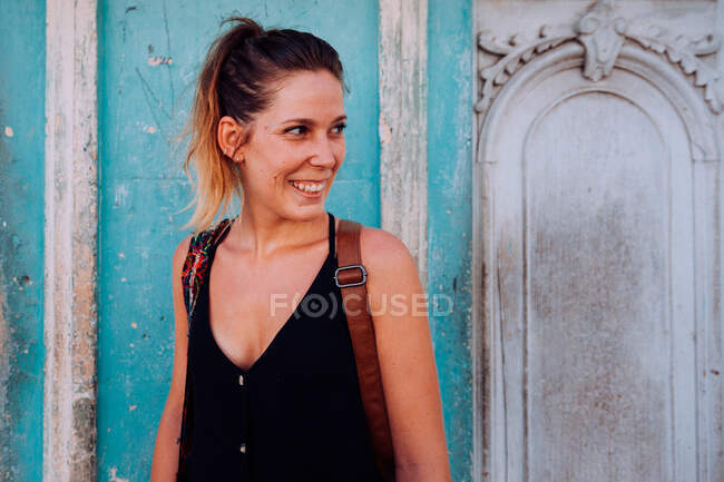 Радісна молода пані у відпустці в чорному повсякденному одязі посміхається і озирається зі стіною старого будинку на Кубі. — стокове фото