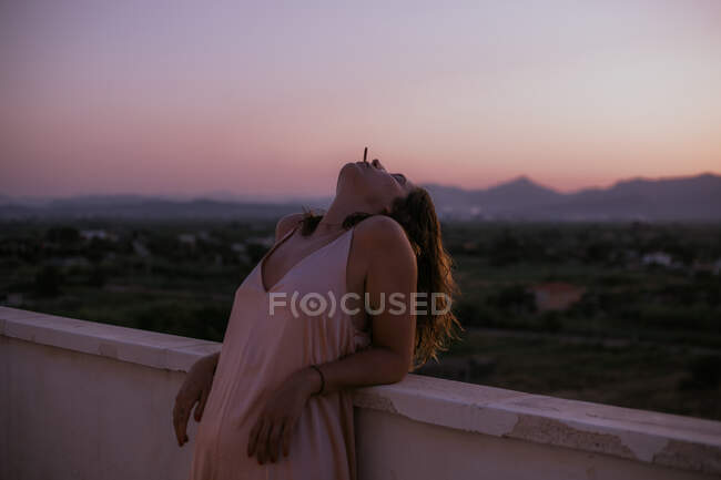Vista lateral da mulher pacífica deitada com os olhos fechados na cerca da varanda e fumar cigarro com o pôr do sol no fundo turvo — Fotografia de Stock