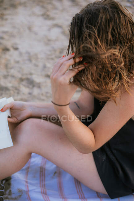 From above of young lady in casual wear with book sitting on sandy beach adjusting hair — Stock Photo
